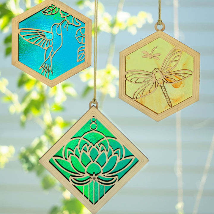 Group of three handcrafted suncatchers with a blurred outdoor background. Suncatchers are a blue hummingbird, yellow dragonfly, and green lotus flower